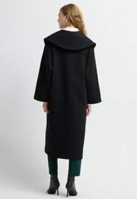 Lange zwarte jas met een brede, gevouwen kraag. Gemaakt van een gladde stof, met rechte mouwen en een ontwerp dat onder de knie valt.