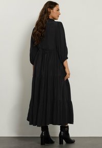 Abito lungo nero con maniche a sbuffo, design a strati e vita cintrata. Presenta un dettaglio con laccio sulla schiena e tessuto testurizzato. Indossato con stivaletti neri.