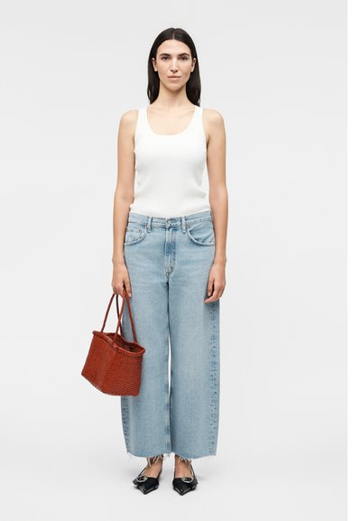 Γυναίκα που φοράει λευκό αμάνικο τοπ, ανοιχτό μπλε τζιν με φαρδιά πόδια, μαύρα μυτερά φλατ παπούτσια, κρατώντας κόκκινη πλεκτή τσάντα, στέκεται μπροστά σε απλό φόντο.