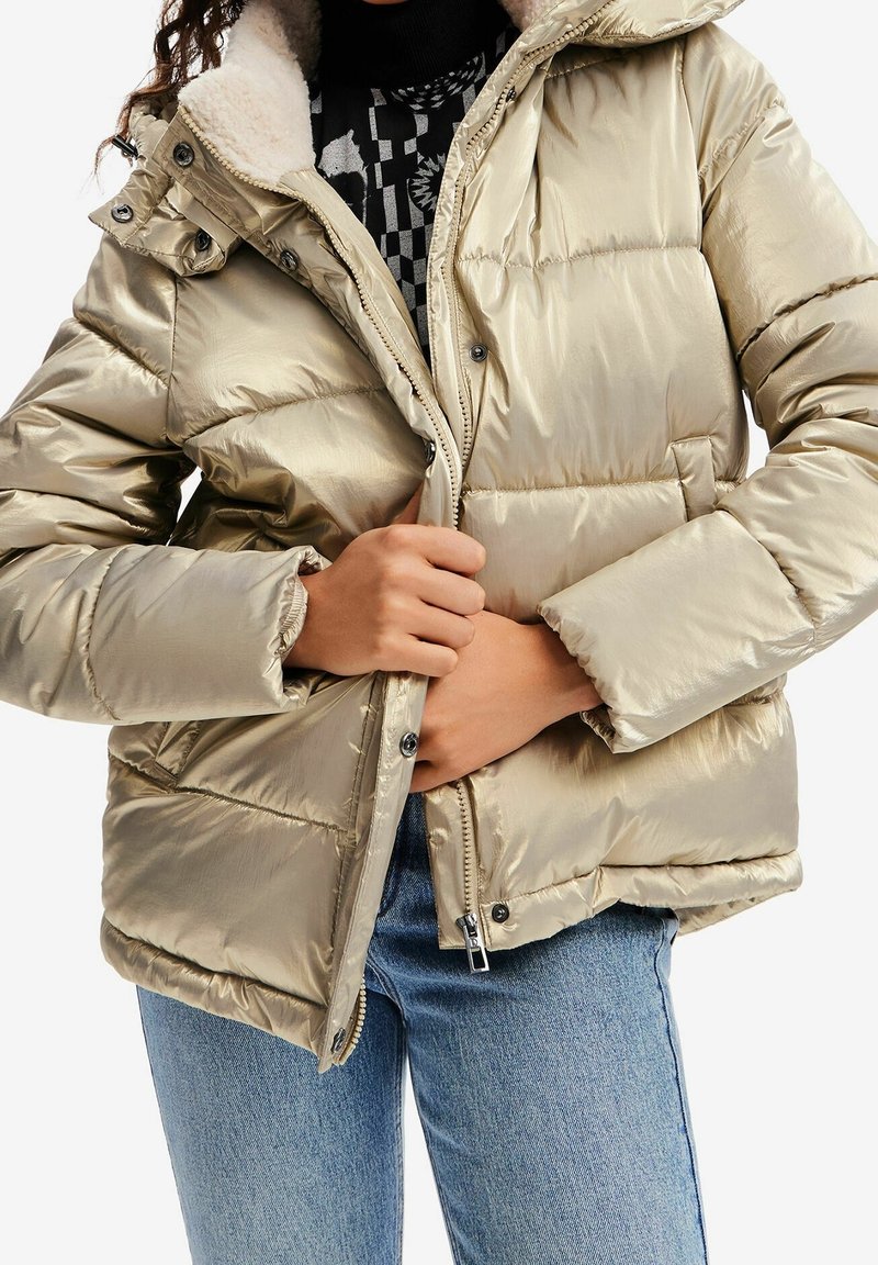 Personne portant une doudoune métallisée beige avec capuche doublée en sherpa, en train de fermer la fermeture éclair, et un jean bleu.
