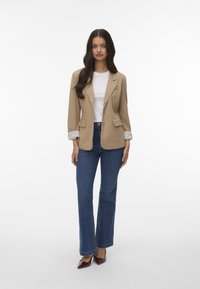 Blazer beige con spalline, top bianco e jeans a zampa d'elefante blu. Il blazer presenta maniche con risvolto e tasche frontali. Scarpe color bordeaux completano l'outfit.