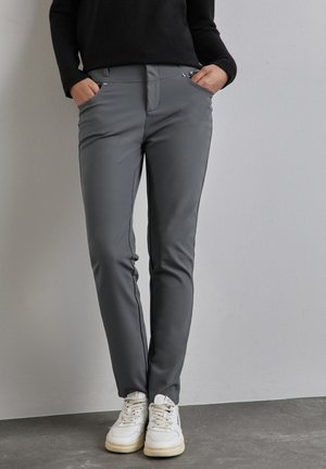 Pantaloni grigi aderenti con una texture liscia, tasche frontali e dettagli in metallo argentato, abbinati a sneakers bianche su una superficie grigia.