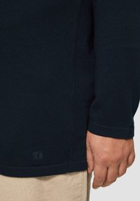 Marineblaues, geripptes Langarmshirt mit abgerundetem Saum und umgeschlagenen Ärmeln, das ein kleines Log etikett an der unteren linken Seite aufweist.