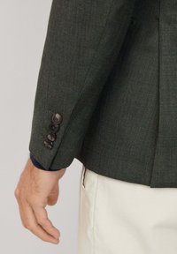 Tmavozelený blejzer so štruktúrovaným tkaním, vybavený štyrmi čiernymi gombíkmi na manžetách a kontrastnou modrou košeľou pod ním.