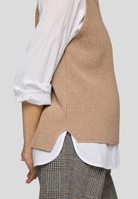 Beige Strickweste mit strukturierter Oberfläche und seitlichem Schlitz, kombiniert mit einem weißen Hemd mit hochgekrempelten Ärmeln und gemusterten grauen Hosen.