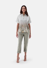 Kurzarm-Hemd mit Knopfleiste und pastellfarbigem Blumenmuster am Saum, kombiniert mit hellgrünen, hochgekrempelten Hosen und beigen Sandalen mit Riemen.