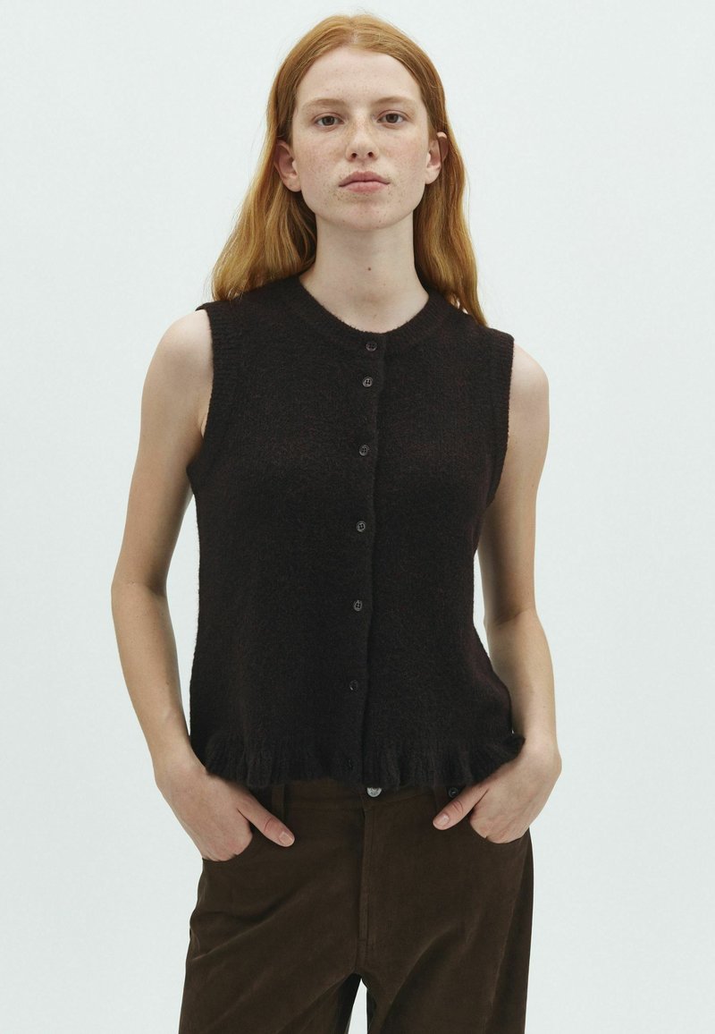 Jeune femme aux longs cheveux roux porte un haut noir sans manches boutonné et un pantalon marron foncé, debout les mains dans les poches sur fond blanc.