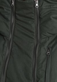 Tmavě zelená prošívaná bunda s třemi černými zipy, dvěma velkými paralelními a jedním menším na pravé kapse.