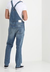 Peto de mezclilla con tirantes ajustables, lavado azul claro, corte relajado y bolsillos delanteros y traseros. Usado sobre una camiseta blanca.