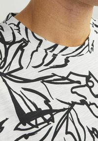 Hvid t-shirt med et sort abstrakt linjemønster. Har en rund halsudskæring og et struktureret stof. Nærbillede af kraven og skulderområdet.