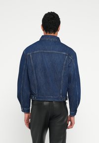 Τζιν μπουφάν σε σκούρο μπλε χρώμα με γιακά, πλαϊνές τσέπες, κουμπιά κλεισίματος και άνετη εφαρμογή, εμφανιζόμενο από πίσω.