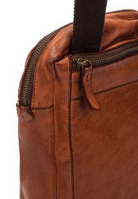 Bruine leren tas met een gestructureerde afwerking, voorzien van een bovenste ritsluiting en een stevige stoffen schouderband. Messing ritsbeslag en stiksels zijn zichtbaar.