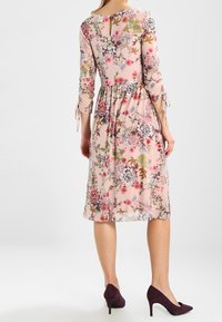 Vestido midi floral en tela rosa translúcida con mangas tres cuartos, puños con lazo y detalle de espalda con abertura. Combinado con tacones morados puntiagudos.