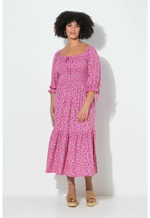 Kvinde med krøllet hår iført en rosa blomsterprint midi-kjole med 3/4 ærmer og tanfarvede platåsandaler, stående mod en lys væg.