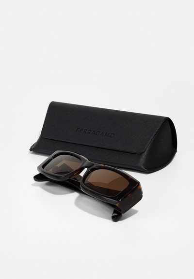 Occhiali da sole tartarugati con lenti marroni, caratterizzati da ampi telai quadrati, posizionati accanto a un astuccio rigido nero con texture, inciso con "Ferragamo."