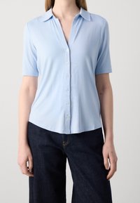 Blusa azzurra a maniche corte, con chiusura a bottoni sul davanti, colletto e texture morbida. Abbinata a pantaloni ampi in denim scuro.
