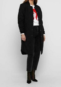 Cappotto lungo trapuntato nero con chiusura a pulsante a pressione, indossato sopra una T-shirt bianca con grafica rossa, abbinato a jeans scuri e stivaletti neri trasparenti.