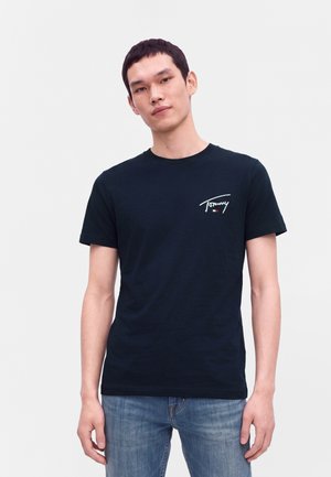 Νέος με κοντά σκούρα μαλλιά, φοράει μαύρο T-shirt με το λογότυπο Tommy και μπλε τζιν, στέκεται μπροστά σε απλό φωτεινό φόντο.