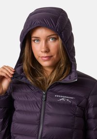 Vrouw met lichte ogen die een donkere paarse pufferjack met rits en capuchon draagt, met het 'Swedemount'-logo erop, terwijl ze de rand van de capuchon vasthoudt.