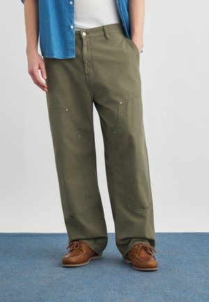 Mann trägt olivgrüne Cargo-Hose, braune Leder-Bootsschuhe, weißes Hemd und blaues Jeanshemd, steht auf blauem Teppich.