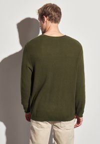 Pull à manches longues vert olive avec un motif de tricot texturé, coupe décontractée, et col rond, associé à un pantalon clair.