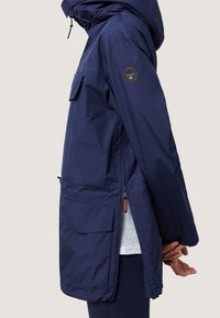 Marineblauwe waterdichte jas met capuchon, zijzakken en ritsdetails. Heeft een badge op de linkermouw en een ontspannen pasvorm.