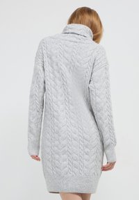 Donna con capelli castano chiaro che indossa un lungo vestito in maglia grigia chiara con un motivo a trecce, mostrata da dietro contro uno sfondo neutro.