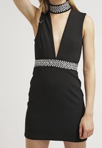 Černé bezramínkové šaty s hlubokým V-výstřihem, ozdobeným krystalovým pásem a odpovídajícím náhrdelníkem. Hladká textura, těsně přiléhající střih.