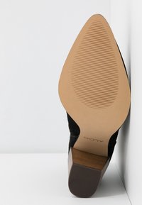 Botte en daim noir à bout pointu avec semelle beige striée et talon bloc en bois, appuyée contre un coin blanc.