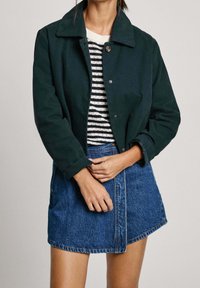 Ciemnozielona skórzana kurtka do połowy talii z wyraźnym kołnierzem, zapięciem na guziki i dżinsową spódnicą, pod którą znajduje się dopasowana bluzka w paski.