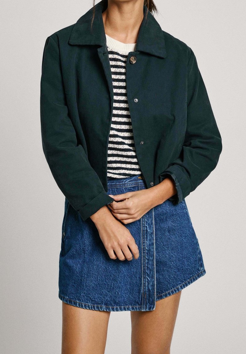 Ciemnozielona skórzana kurtka do połowy talii z wyraźnym kołnierzem, zapięciem na guziki i dżinsową spódnicą, pod którą znajduje się dopasowana bluzka w paski.