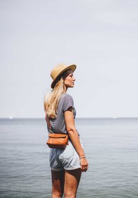 Hellbeige Strohhut, grau gestreiftes Hemd, Jeansshorts und orangefarbene Leder-Umhängetasche. Beruhigendes Meer im Hintergrund mit klarem Himmel.