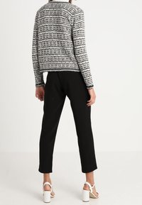 Svart och vit mönstrad jacka med en texturerad yta, bärs med svarta byxor och vita klacksandaler, sett från en bakvy.