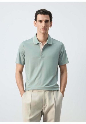 Hombre de cabello oscuro con un polo verde claro de manga corta y pantalones beige, de pie con las manos en los bolsillos frente a un fondo liso.
