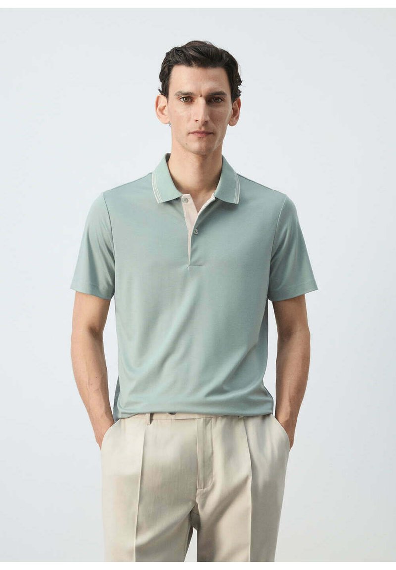 Homme aux cheveux foncés portant un polo vert clair à manches courtes et un pantalon beige, debout les mains dans les poches devant un fond uni.