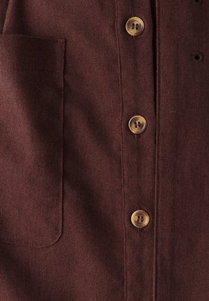 Camisa marrón de botones con una tela texturizada, que presenta cuatro botones de color claro y un bolsillo en el pecho del lado izquierdo.