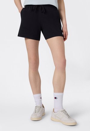 Persona con pantalones cortos negros con cordón, calcetines blancos con el logotipo de Champion y zapatillas beige, de pie sobre un fondo blanco liso.