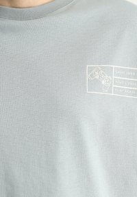 Camiseta de algodón gris con un gráfico impreso de un mando de videojuego y el texto "GAME OVER", "NEXT LEVEL" y "PLAY AGAIN" en blanco.