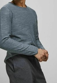 Personne portant une chemise grise texturée à manches longues et un pantalon gris foncé, debout les mains jointes devant un fond uni.