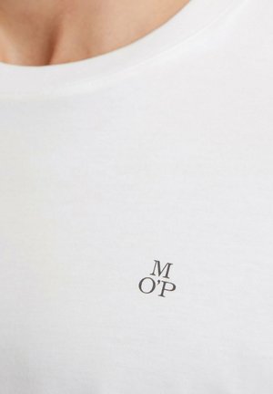 Primer plano de una persona que lleva una camisa blanca con pequeñas letras negras "M O'P" impresas cerca del área del pecho.