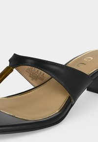 Μαύρο πέδιλο με τακούνι, φαρδιά δερμάτινη λωρίδα πάνω από το πόδι και ξύλινη υφή στη λωρίδα ανάμεσα στα δάχτυλα, μπεζ πάτος με χρυσό περίγραμμα και ετικέτα μάρκας.