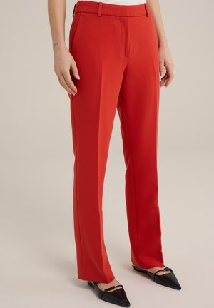 Kvinde iført skræddersyede, klare røde bukser med forreste folder og sorte spidse flade sko med ankelstropper, stående mod en ensfarvet baggrund.