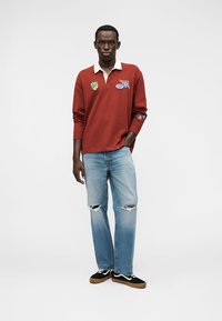 Κόκκινο πόλο με μακριά μανίκια, λευκό γιακά και επιθέματα, συνδυασμένο με ανοιχτό μπλε τζιν με σκισίματα και μαύρα αθλητικά παπούτσια με καφέ σόλες.