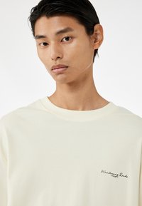 Hellgelbes Baumwollsweatshirt mit Rundhalskragen und kleinem schwarzen gesticktem Logo auf der linken Brust. Glatte Textur.