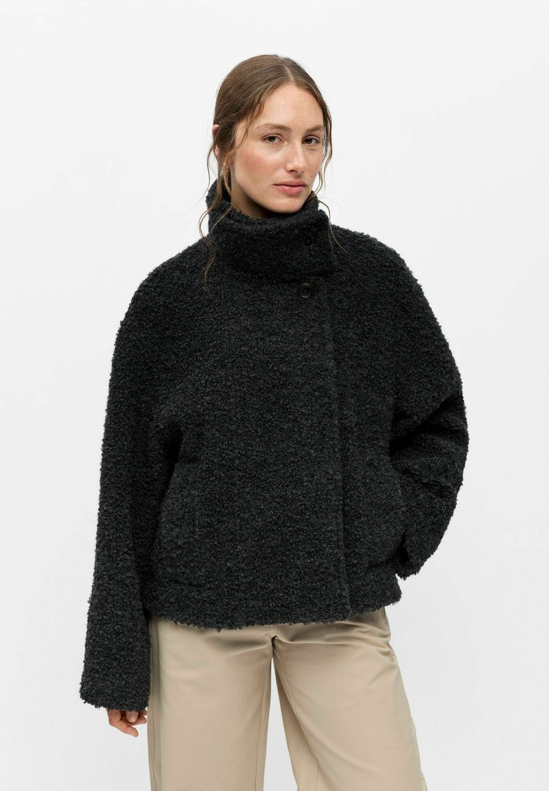 Femme portant une veste noire texturée à col haut avec manches longues et un pantalon beige, debout les mains dans les poches devant un fond uni.