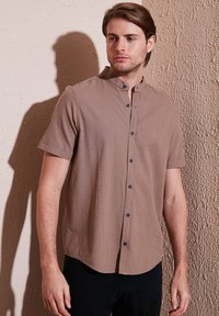 Korte mouwen, bruine shirt met een mandarin-kraag, voorzien van zwarte knopen en een gestructureerde stof. Draagt met donkere broek tegen een lichte muur.