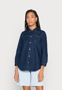 Camicia in denim blu scuro con maniche lunghe, chiusura a bottoni e colletto. Presenta cuciture chiare e un leggero orlo arrotondato. Indossata con pantaloni corti chiari.