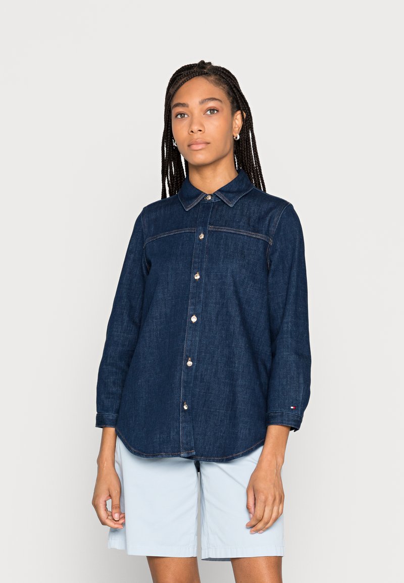 Camicia in denim blu scuro con maniche lunghe, chiusura a bottoni e colletto. Presenta cuciture chiare e un leggero orlo arrotondato. Indossata con pantaloni corti chiari.