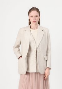 Beige linblazer med en enkelt knapp, notch-lapper og to frontlommer, båret over en lyseblå topp og en plisserte rosa skjørt.