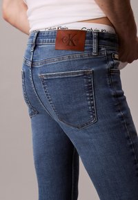 Férfi kék Calvin Klein farmernadrágban, látható barna bőr márkajelzéssel és fehér Calvin Klein fehérnemű derékpánttal.
